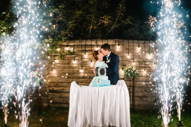 Photo of Bride and a groom is kissing near rustic wedding cake with firework in night on wedding banquet outdoors.