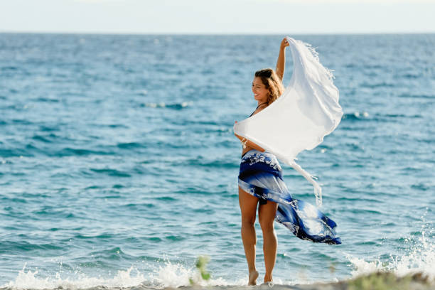 figlarna kobieta bawiąc się szalem w letni dzień nad morzem. - women sarong beach white zdjęcia i obrazy z banku zdjęć