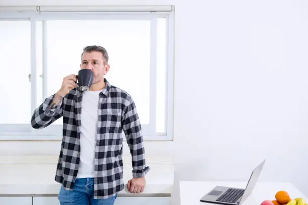 Picture of mature man drinking a cup of coffee while standing near a laptop in the kitchen