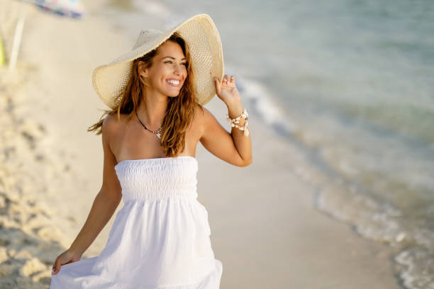 Carefree woman going for a walk on the beach in summer day. beautiful happy woman wearing sundress and straw hat having fun while walking on the shore. sundress stock pictures, royalty-free photos & images