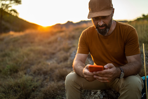 Mature man with cap sitting on stone and using smart phone in nature