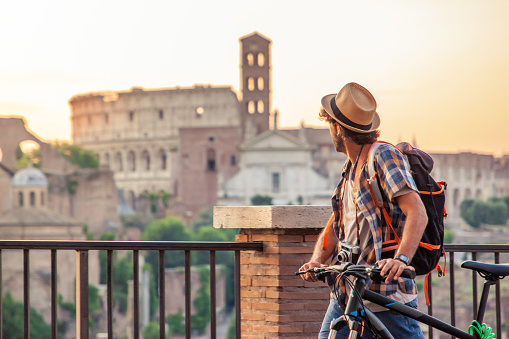 Young man tourist with backpack and bike walking at Roman Forum at sunrise. Historical imperial Foro Romano in Rome, Italy from panoramic point of view.