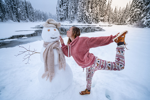 Christmas time; Young woman kissing snowman in snowy mountain landscape; having fun in winter vacations