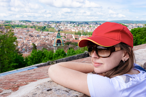 Young girl looking at Cityscape from Citadel of Besancon at Bourgogne Franche-Comte region, France. Lady wearing sunglasses and red baseball cap at Town roofs seen from French Castle in Burgundy.