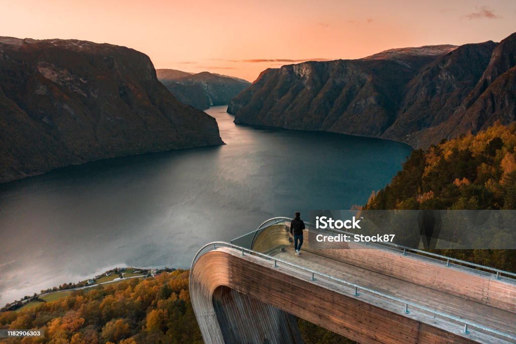 A Tourist Views Aurlandsfjord From the Stegastein Lookout in Norway at Sunset A tourist takes in the view from the Stegastein viewpoint overlooking the Aurlandsfjord at sunset with a beautiful sunset Norway Stock Photo
