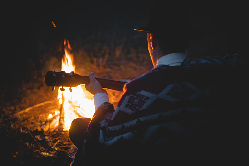 Man in traditional native american poncho and hat plays the guitar by the fire in the wood