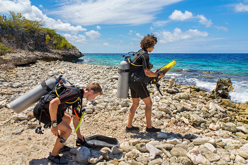 Two young caucasian divers on the beach walking to the sea. These sporty men are almost ready to dive in the blue sea where many beautiful fishes can be seen under water. This shore is filled with boulders.