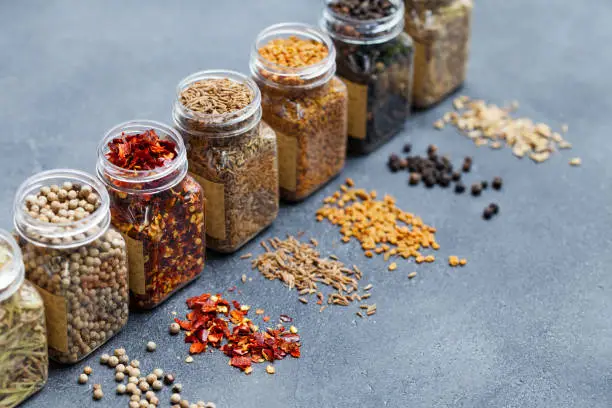 Photo of Assortments of spices, white pepper, chili flakes, lemongrass, coriander and cumin seeds in jars on grey stone background. Copy space.