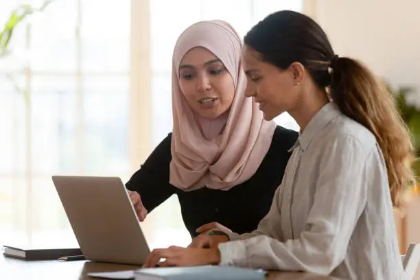 Photo of Asian muslim female mentor teaching caucasian intern explaining computer work