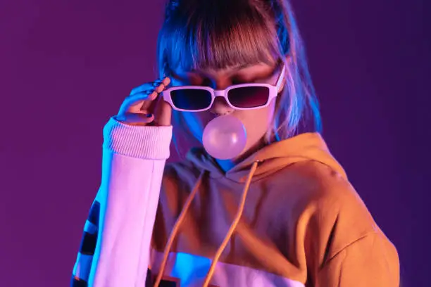 Photo of Pretty young 20s fashion teen girl model wear stylish glasses hoodie blowing bubble gum standing at purple violet studio wall background, igen teenager in trendy night party glow 80s 90s concept