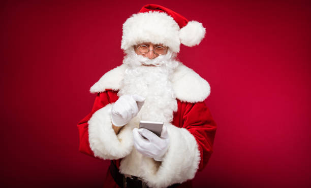 istock Checking tasks. Santa Claus in round-shaped glasses is posing with a smartphone in his left hand and with his right hand pointing at something on the screen. 1182958255