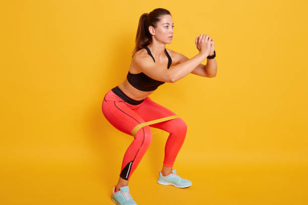 tiro de estudio de mujer deportiva en cuclillas, haciendo sentarse con la banda de resistencia. foto de mujer caucásica en ropa deportiva de moda aislada sobre fondo amarillo. concepto de fuerza y motivación. - sit ups fotografías e imágenes de stock