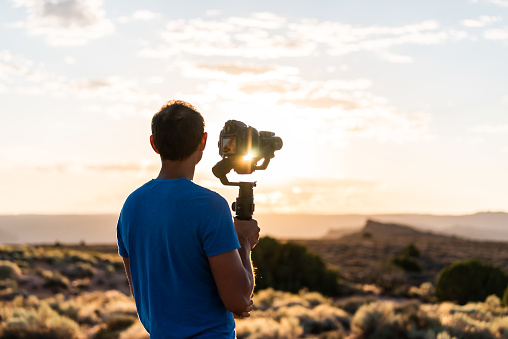 Man holding gimbal camera stabilizer filming sunrise with sunburst flare through equipment in Arches National Park, Utah at Fiery Furnace Viewpoint