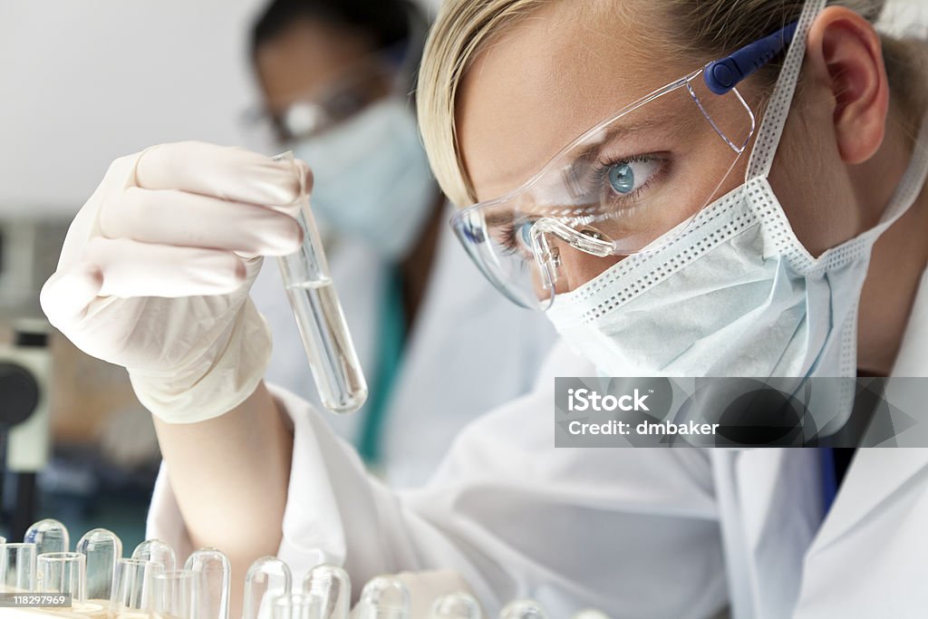 Female Scientific Research Team With Clear Solution In Laboratory A blond medical or scientific researcher or doctor using looking at a clear solution in a laboratory with her Asian female colleague out of focus behind her. Test Tube Stock Photo