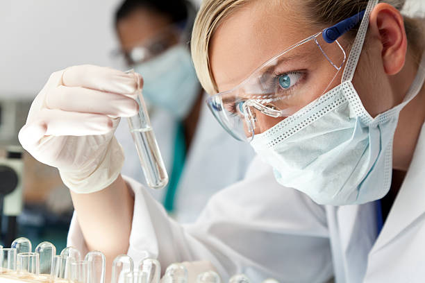 Female Scientific Research Team With Clear Solution In Laboratory A blond medical or scientific researcher or doctor using looking at a clear solution in a laboratory with her Asian female colleague out of focus behind her. science research stock pictures, royalty-free photos & images