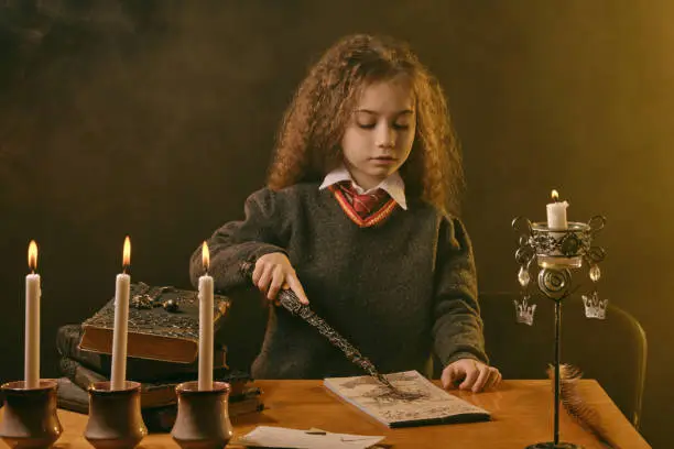 Photo of Little witch dressed in dark clothes sitting at the table against a black smoky background. There are magic wand, books with spells, candles on it