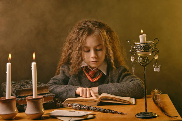 Little witch dressed in dark clothes sitting at the table against black smoky background and reading a book. Close-up portrait Portrait of a cute little witch girl with magnificent long brown hair dressed in a navy blue jumper, white shirt and red tie. She is reading a book with spells while sitting at the table against a black smoky studio background. There are magic wand, candles in candlesticks on it. The concept of magic, wizardry, miracles. Close-up. wizard photos stock pictures, royalty-free photos & images