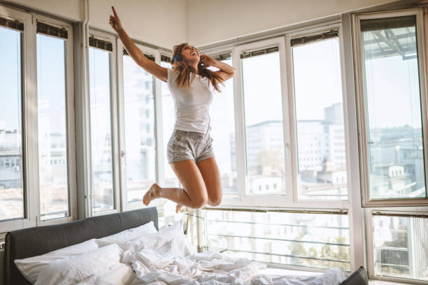 Waking up routine Charming young woman at home listening music and jumping at bed air jump stock pictures, royalty-free photos & images
