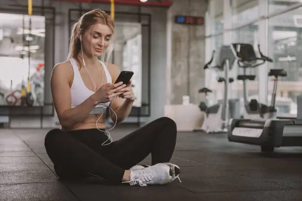 Happy healthy young sportswoman sitting on the floor at the gym, using her smart phone, copy space. Fitness woman resting at sport studio after exercising, browsing online on her phone