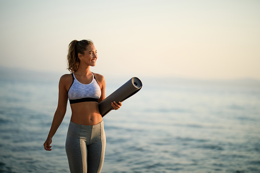 Young happy sportswoman carrying exercise mat while walking by the sea in the morning. Copy space.
