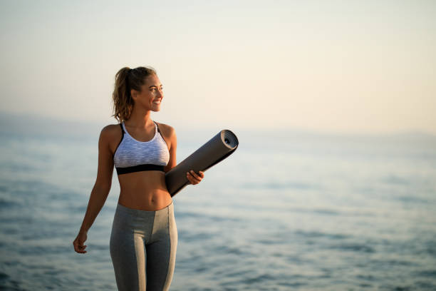 Happy athletic woman walking at the beach. Young happy sportswoman carrying exercise mat while walking by the sea in the morning. Copy space. sports bra stock pictures, royalty-free photos & images