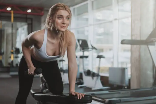 Happy beautiful female athlete smiling, doing triceps exercise at sport studio, copy space. Lovely fitness woman enjoying her morning workout at the gym, lifting dumbbells