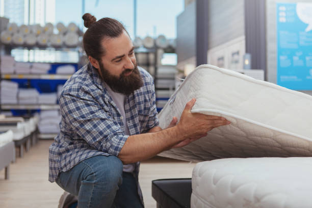 Mature man buying orthopedic bed at furniture store Household shopping concept. Mature bearded man examining orthopedic mattress at furnishings store. Handsome male customer choosing new bed at furniture shop mattress stock pictures, royalty-free photos & images