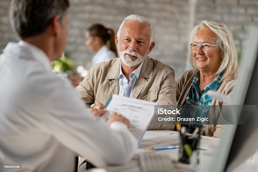 Happy senior couple going through medical insurance paperwork with a doctor. Happy mature couple informing themselves about health insurance while talking to a doctor at clinic. Medicare Stock Photo