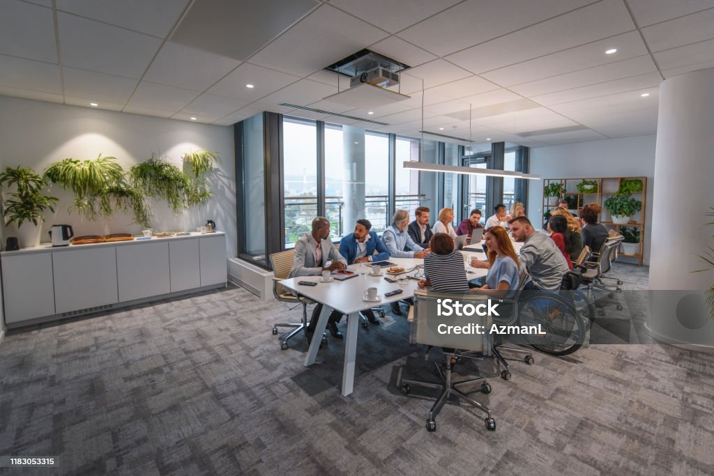 Executive Team Sitting at Conference Table in Board Room Distant view of management team sitting at conference table and talking with each other prior to start of weekly meeting. Organized Group Stock Photo