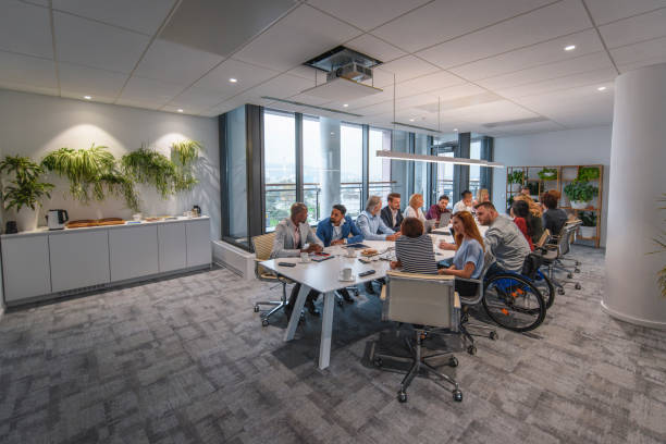 Executive Team Sitting at Conference Table in Board Room Distant view of management team sitting at conference table and talking with each other prior to start of weekly meeting. persons with disabilities photos stock pictures, royalty-free photos & images