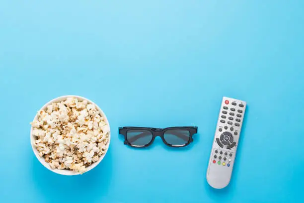 Bowl with popcorn, Imax glasses, remote control for TV on a blue background. Concept home theater, novelties of cinema, leisure. Flat lay, top view.