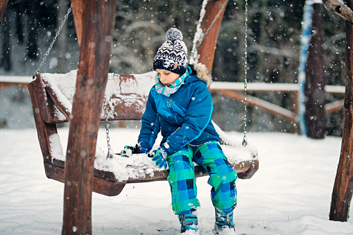 Portrait of a cute boy on winter day. The boy is playing with snow on swing in the back yard\nNikon D850