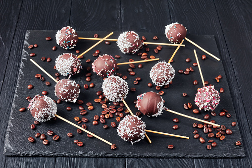 homemade chocolate cake pops sprinkled with crushed candies and coconut sprinkles on a black stone tray with coffee grains, close-up
