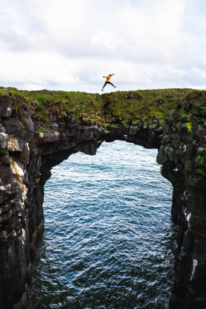 istock Tourist jumps over a natural rock bridge in Arnarstapi, Iceland 1183082971