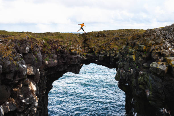 tourist springt über eine natürliche felsbrücke in arnarstapi, island - snaefellsnes stock-fotos und bilder