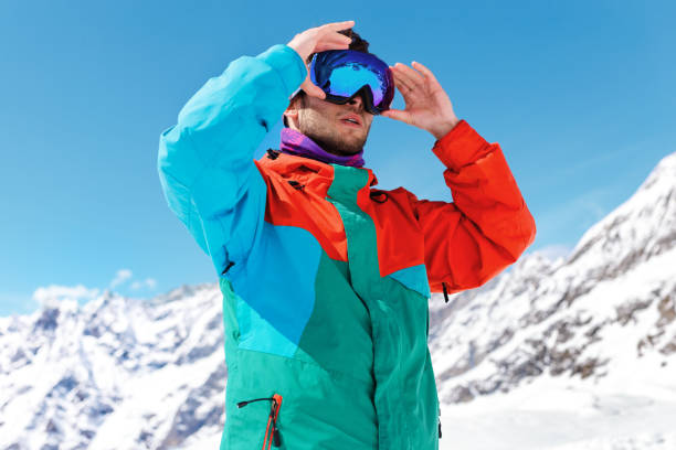 Young skier, wearing ski glasses in the mountains and colorful clothes, sitting over clear sky and sunny day background. stock photo