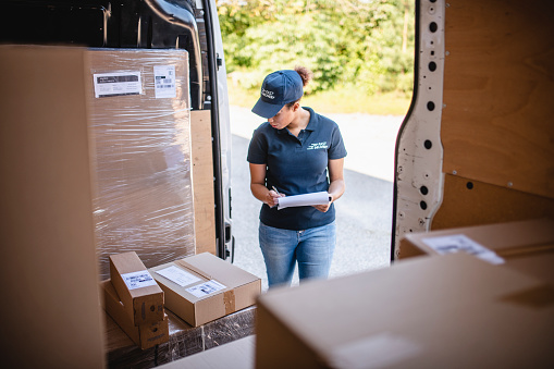 Interior view of mixed race female delivery expert checking packages loaded on van against loading list.