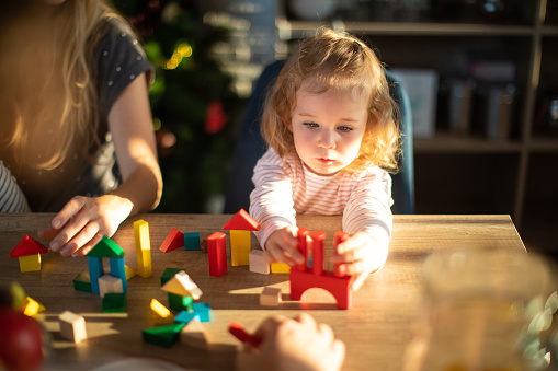 Blond hair toddler girl playing with toy blocks at home, sitting at dining table, unrecognizable people