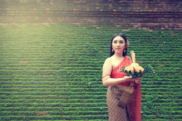 Loy Krathong Festival, Beautiful young Thai woman in traditional dress looking up holding Krathong made by banan leafs candle incense yellow flowers on green grass background.