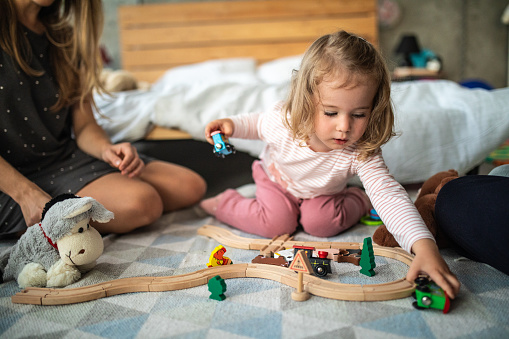 Cute two years old girl sitting on floor and having fun with wooden train toy, parents sitting next to her