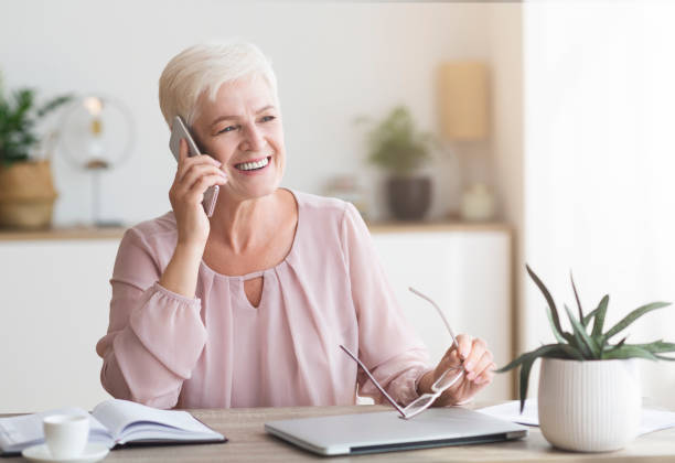 istock Smiling senior woman talking by phone at workdesk at home 1183102021