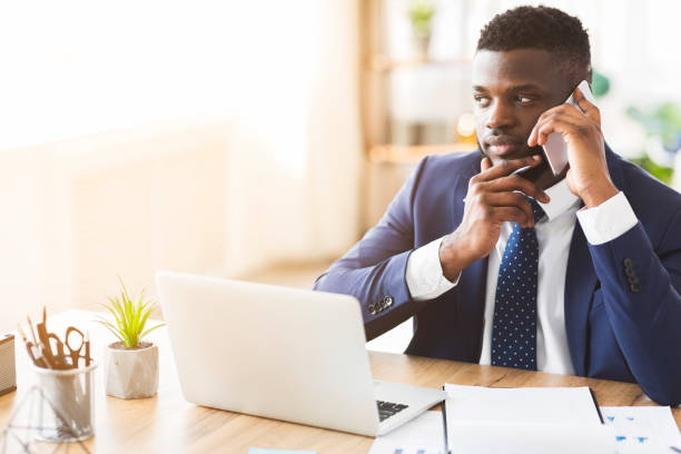 Young african entrepreneur having pleasant conversation with business partner by phone, office interior, copy space