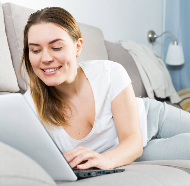 istock Cheerful girl is lying on sofa and playful posing with laptop 1183104826