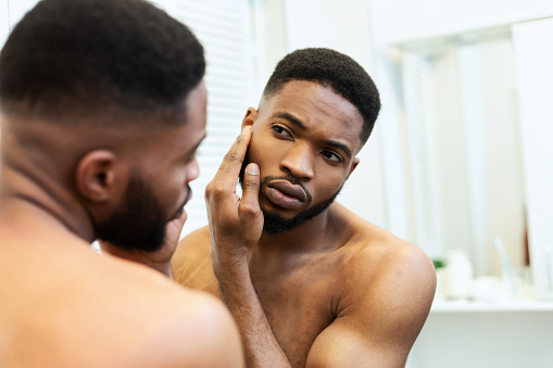 Acne problem. Young african american guy checking his face skin in mirror, free space