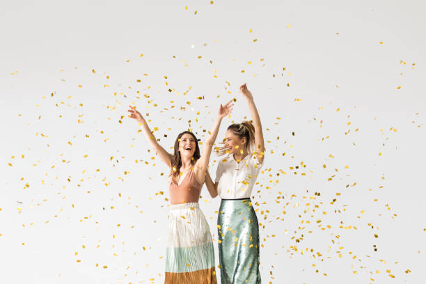 Studio shot of two beautiful stylish young women smiling and dancing under confetti.