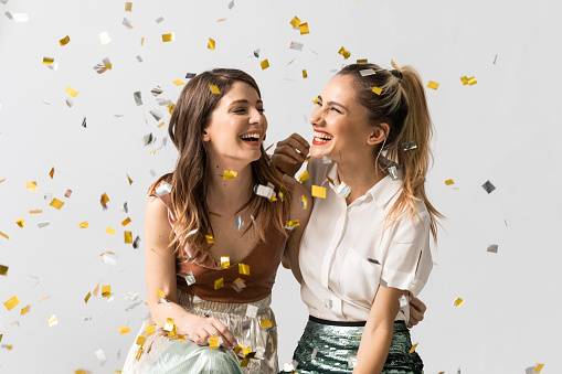 Portrait of two beautiful laughing young women feeling happy together celebrating under confetti.