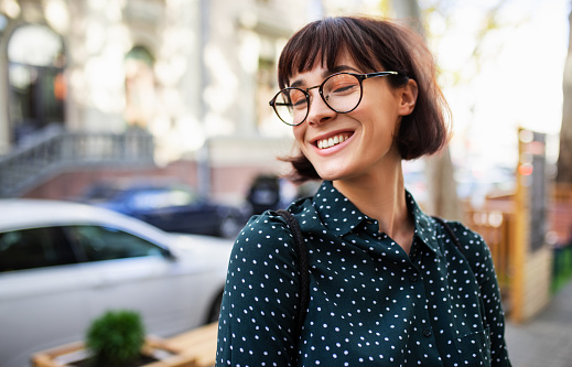 Outdoor portrait of beautiful young woman wearing green shirt with white dots and transparent eyeglasses, smiling and laughing. Young female student posing in the city street.