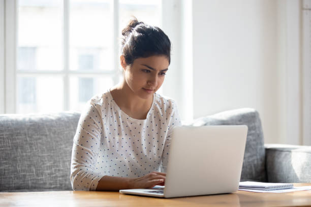 donna indiana concentrata che usa il laptop a casa, guardando lo schermo - select focus foto e immagini stock