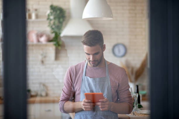 istock Dark-haired man wearing striped apron reading recipe on tablet 1183154224