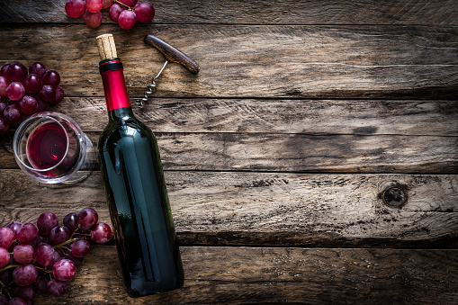 Top view of a rustic table with a composition of red wine bottle, wineglass, corkscrew and grapes  arranged at the left border leaving useful copy space for text and/or logo at the right. Predominant colors are red and brown. XXXL 42Mp studio photo taken with Sony A7rii and Sony FE 90mm f2.8 macro G OSS lens
